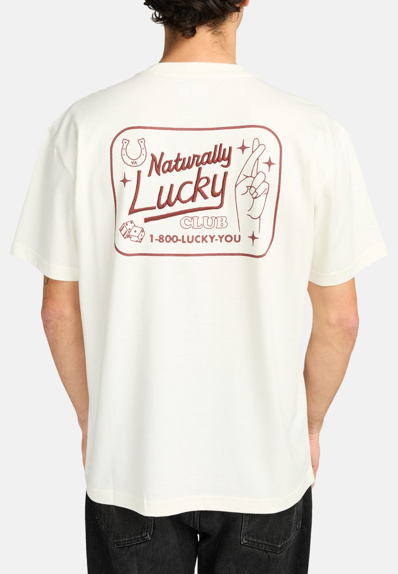 Cream-colored t-shirt with a reddish graphic on the back featuring the text "Naturally Lucky Club," dice, and crossed fingers design.