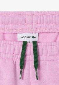 Shorts rose clair avec une ceinture texturée, dotés de cordons de serrage verts. Étiquette emblématique Lacoste visible sur la ceinture.