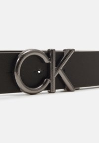 Black textured leather belt featuring a glossy black metal buckle with embossed "CK" logo. The buckle has a circular and angular design.
