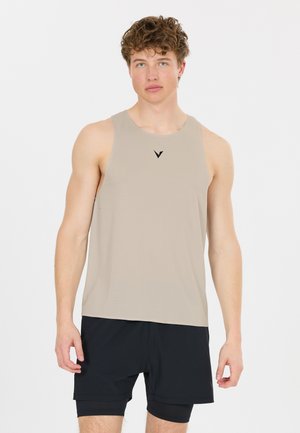 Junger Mann mit lockigem Haar, der ein beiges ärmelloses Sportshirt und schwarze Shorts trägt, steht vor einem weißen, schlichten Hintergrund.