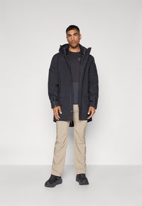 Parka negra impermeable con capucha, dos bolsillos frontales, puños ajustables y cierre con cremallera. Llevada sobre una camiseta oscura y pantalones beige.