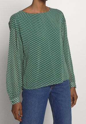 Green long-sleeve blouse with white diamond pattern, paired with blue jeans, worn by a person with medium-dark skin tone.