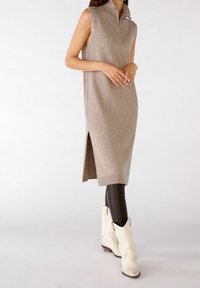 Robe sans manches en tricot couleur marron clair avec un col à demi-zip, fentes latérales, associée à des leggings noirs brillants et des bottes blanches de style western.