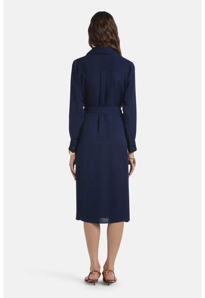 Žena stojaca chrbtom, oblečená v tmavomodrých midi šatách s dlhým rukávom a opaskom, so hnedými sandálmi na nízkom podpätku.