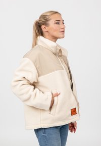 Beżowa i kremowa polarowa kurtka z kołnierzem na stojąco, kieszeniami bocznymi i pomarańczową metką. Miękka tekstura, luźny krój i ciepły design.