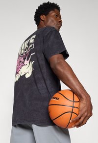 Čierne tričko s tie-dye dizajnom a prednou grafikou. Krátke rukávy, voľný strih. Drží oranžový basketbal s čiernymi detailmi.