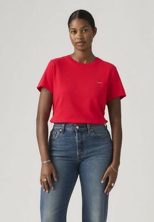 Vrouw met donker haar in een rood T-shirt met korte mouwen en blauwe jeans met hoge taille, staand tegen een effen lichte achtergrond.