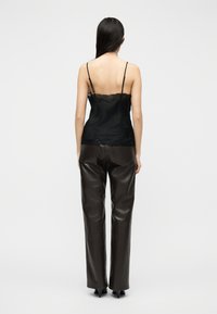 Γυναίκα με μακριά σκούρα μαλλιά, που φοράει μαύρο καμισόλι με δαντέλα και σκούρο καφέ δερμάτινο παντελόνι, στέκεται με την πλάτη γυρισμένη σε λευκό φόντο.