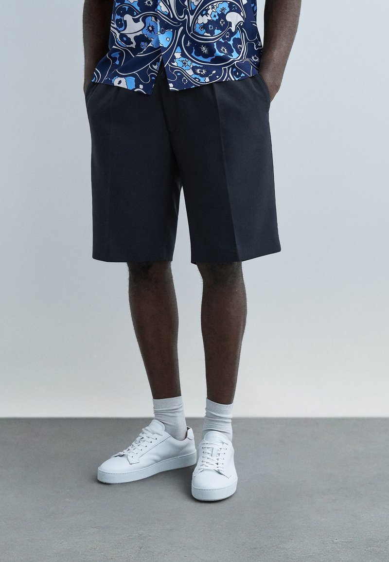 Homme portant un short bleu marine, des chaussettes blanches, des baskets blanches et une chemise bleue à motifs, les mains dans les poches.