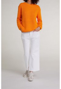 Maglione arancione lavorato a maglia con motivo a rilievo, abbinato a pantaloni bianchi a gamba larga e sneakers grigie. Il maglione ha maniche lunghe e scollo rotondo.