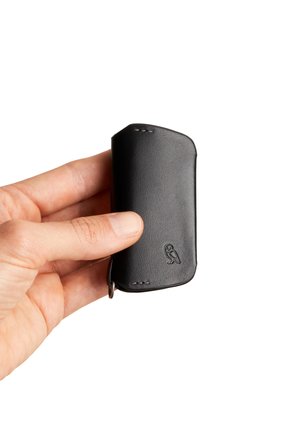 Porte-clés en cuir noir avec bords arrondis, présentant un logo embossé, des détails cousus à la main et un attachement à anneau métallique.