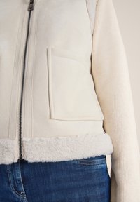 Veste en daim synthétique beige avec des manches en tricot, fermeture éclair verticale et une poche avant. Le bas présente un bord doux et duveteux.