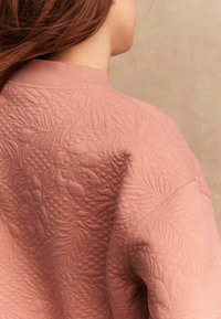 Femeie cu păr lung brunet, îmbrăcată într-o jachetă roz matlasată cu modele florale și frunze, văzută din spate, pe un fundal bej.