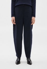 Άτομο που φοράει ραμμένα παντελόνια σε ναυτικό μπλε χρώμα, μαύρα δερμάτινα μποτάκια μέχρι τον αστράγαλο και σκουρό μπλε πουλόβερ, στέκεται μπροστά σε λευκ�ό μονόχρωμο φόντο.