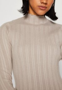 Maglione a collo alto beige a coste, realizzato in materiale morbido, con un collo alto e un tessuto leggermente testurizzato con righe verticali.