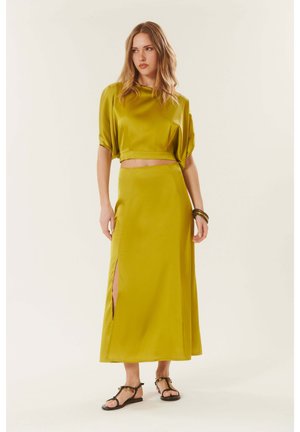 Femme portant un crop top en satin jaune moutarde et une longue jupe fendue sur le côté, assortis à des sandales noires et des bracelets, debout sur un fond uni.