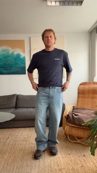 Ναυτική μπλούζα με λογότυπο, ανοιχτό μπλε τζιν παντελόνι, χαλαρή εφαρμογή, φορεμένα με σκούρα αθλητικά παπούτσια. Εσωτερικός χώρος με καναπέ και έργα τέχνης.