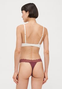 Bralette blanche avec des bretelles réglables et une fermeture à crochet ; associée à un string bordeau avec dentelle et bordure festonnée, offrant une couverture minimale.