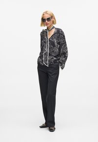 Fekete blúz fehér levélmintával és megkötős részlettel, fekete nadrággal és lapos cipővel párosítva. A modell napszemüveget visel.