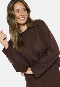 Pruunist kudumist mütsist, millel on ümmargune kaelus ja pikad varrukad, tekstuuriga kangas ja peene sära. Näha on sobiv vöökoht.