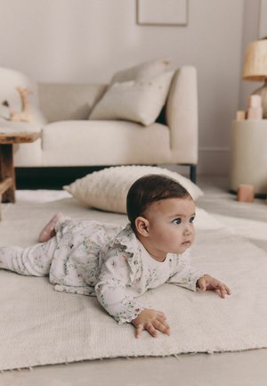 Dziecięcy strój w kwiatowy wzór z falbanami na rękawach, jasna tkanina. Niemowlę leży na teksturowanym beżowym dywanie w miękkim pokoju o neutralnych tonach.