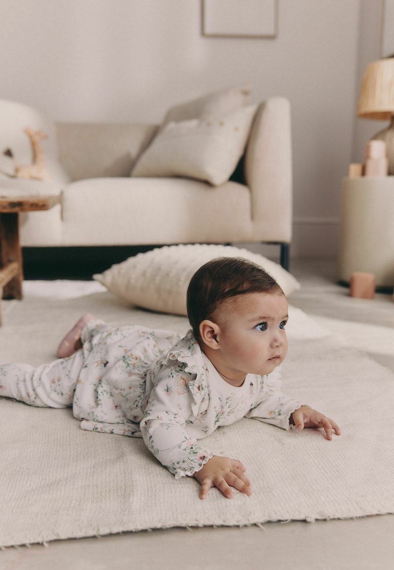 Conjunto para bebé con estampado floral y mangas con volantes, de tela clara. Bebé acostado sobre una alfombra beige texturizada en una habitación suave con tonos neutros.