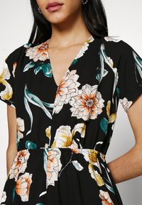 Combinaison à fleurs avec un fond noir, des fleurs vibrantes orange et blanches, un décolleté en V profond, des manches courtes et une taille élastique pour un bon ajustement.