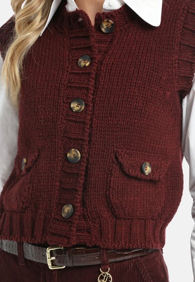 Kvinna som bär en vinröd stickad väst med knappar och fickor över en vit skjorta med krage, ihopkopplad med matchande byxor och ett mörkt bälte.