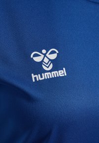 Niebieska sportowa koszulka wykonana z gładkiej tkaniny, z białym logo przedstawiającym stylizowaną pszczołę oraz nazwę marki "hummel" wyraźnie widoczną.
