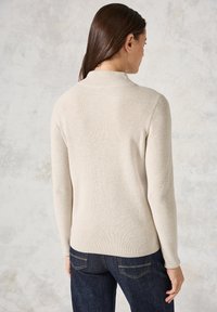 Beige geripptes Sweatshirt mit langen Ärmeln, hohem Kragen, tailliertem Design und glatter Textur, kombiniert mit dunkelblauen Jeans.