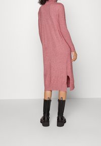Robe longue en tricot rose avec un col montant, présentant une fente latérale et des manches longues. Portée avec des bottes noires jusqu'aux genoux. Texture lisse.