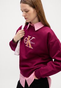 Bordový sveter s vypuklým logom "CK", vrstvený nad ružovou a bielou prúžkovanou košou s golierom. Hladká tkanina, voľný strih a dlhé rukávy.