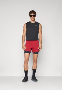 Black sleeveless top, red shorts with navy accents, black socks, and black shoes. Model stands against a plain background.