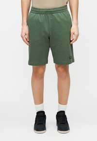 Rohelised puuvillased lühikesed püksid elastse vöökohaga, küljetaskutega ja tagasihoidliku brändinguga alumises vasakus servas. Sobitatud mustade tossude ja valgete sokkidega.