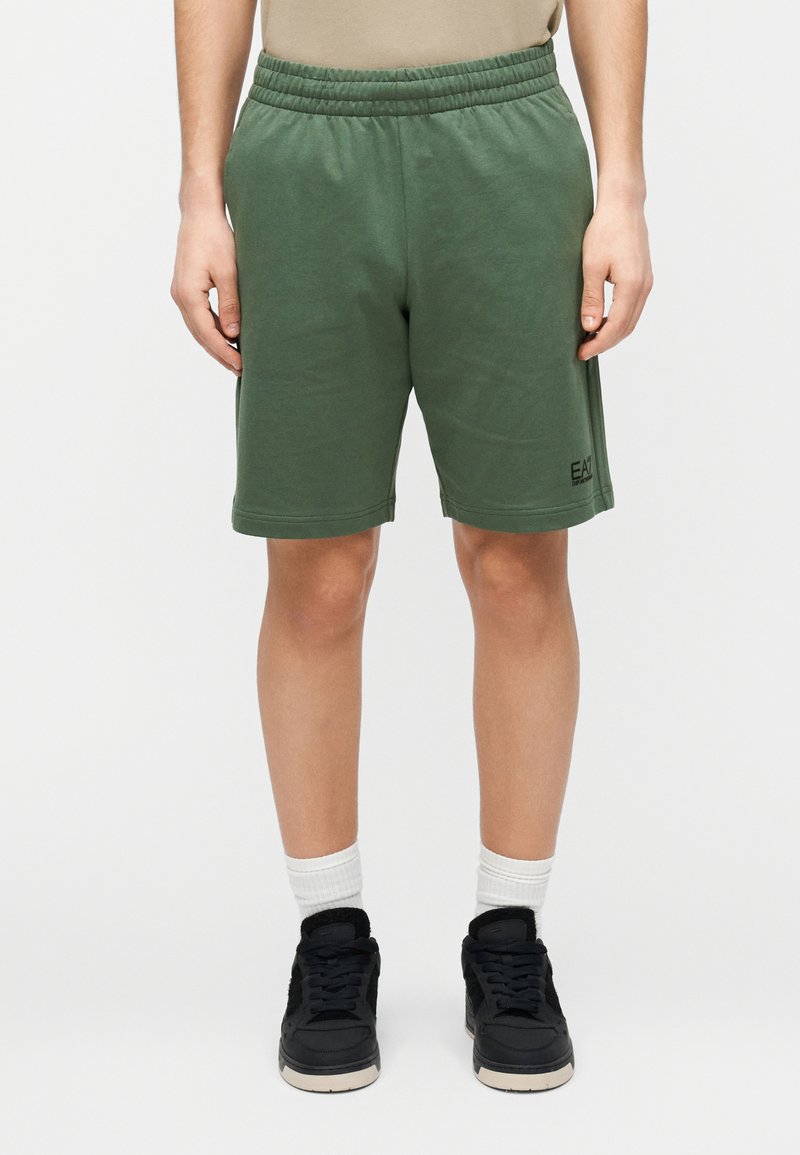 Πράσινα βαμβακερά σορτς με ελαστική μέση, πλαϊνές τσέπες και διακριτικό λογότυπο στο κάτω αριστερό μέρος. Συνδυασμένα με μαύρα αθλητικά παπούτσια και άσπρες κάλτσες.