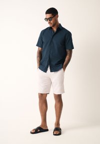 Navy T-shirt met korte mouwen, witte korte broek en zwarte sandalen. Het shirt heeft een knoopsluiting; de korte broek heeft een losse pasvorm en een gladde stof.