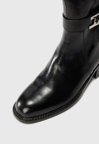 Bottine en cuir noir à bout arrondi, texture lisse et accent de boucle métallique décoratif sur la sangle. Talon bloc bas.