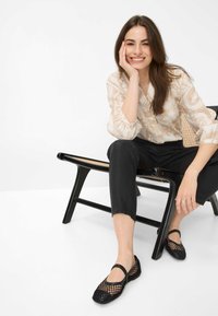 Zwarte geweven mesh ballerina's met een ronde neus en enkelbandje, gecombineerd met zwarte broek en een beige blouse met patroon, zittend op een houten stoel.
