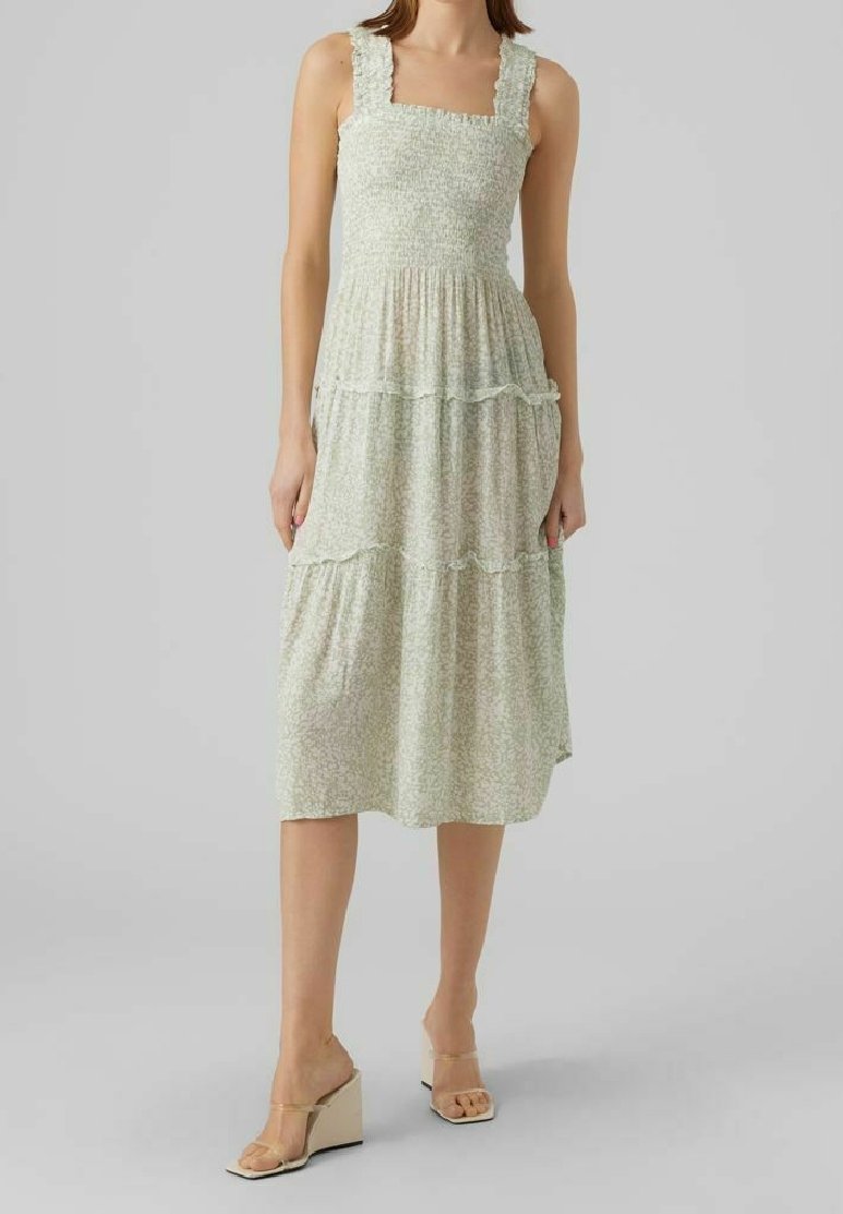 Femme portant une robe midi sans manches, de couleur claire, avec un col carré et une jupe à volants, associée à des sandales compensées beige.