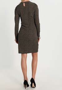 Robe à manches longues et à motifs en vert foncé avec un dos en trou de serrure, silhouette ajustée et accents de boutons aux poignets. Portée avec des talons noirs.