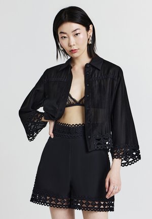 Young woman wearing black lace-trimmed shorts, matching bralette, and sheer button-up shirt with lace details on sleeves and hem.