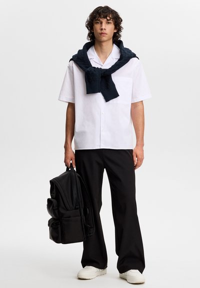 Młody mężczyzna ubrany w białą koszulę z krótkim rękawem, czarne spodnie, białe trampki, ciemny sweter zarzucony na ramiona, trzymający czarny plecak.