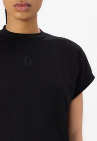 Personne portant un t-shirt noir avec un petit logo smiley en relief sur la poitrine, visible des épaules à mi-torse.