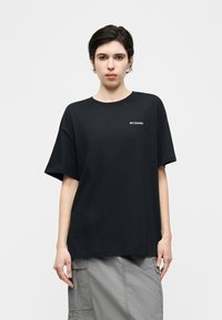 Άτομο που φοράει μαύρη μπλούζα Columbia και γκρι cargo φούστα, όρθιο μπροστά σε απλό λευκό φόντο, κοιτάζοντας μπροστά.