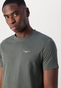 Tricou scurt cu mânecă, de culoare verde închis, realizat din bumbac. Are guler rotund și un mic text al logo-ului alb pe pieptul stâng. Textură netedă.