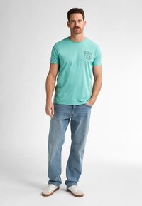 Tyrkysové tričko s krátkým rukávem a předním potiskem, v kombinaci se světle modrými džíny a bílými teniskami s hnědými detaily.