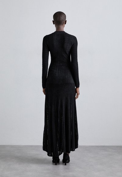 Person med kort hår iført en tætsiddende, lang sort strikket kjole og hæle, stående op ad en simpel lys grå væg og betongulv.