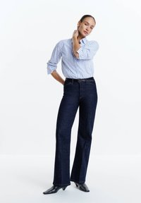 Vrouw staat met hand aan de nek, draagt een wit-blauw gestreept shirt, donkerblauwe spijkerbroek, zwarte riem en zwarte laarzen met hak.