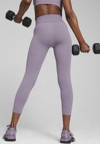 Šviesiai purpurinės, prie kūno prigludusios legging'ų kelnės su aukštu liemeniu ir tekstūruotais akcentais, derinamos su juodais hanteliais ir daugiaspalviais sportiniais bateliais.