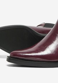 Par de botas de tobillo de cuero rojo oscuro brillante con paneles laterales elásticos negros y suelas de goma texturizada negras.